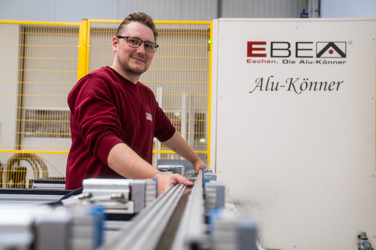 Mitarbeiter im roten EBE-Pullover spannt ein Aluminiumprofil an einer Maschine mit der Aufschrift „Alu-Könner“ ein