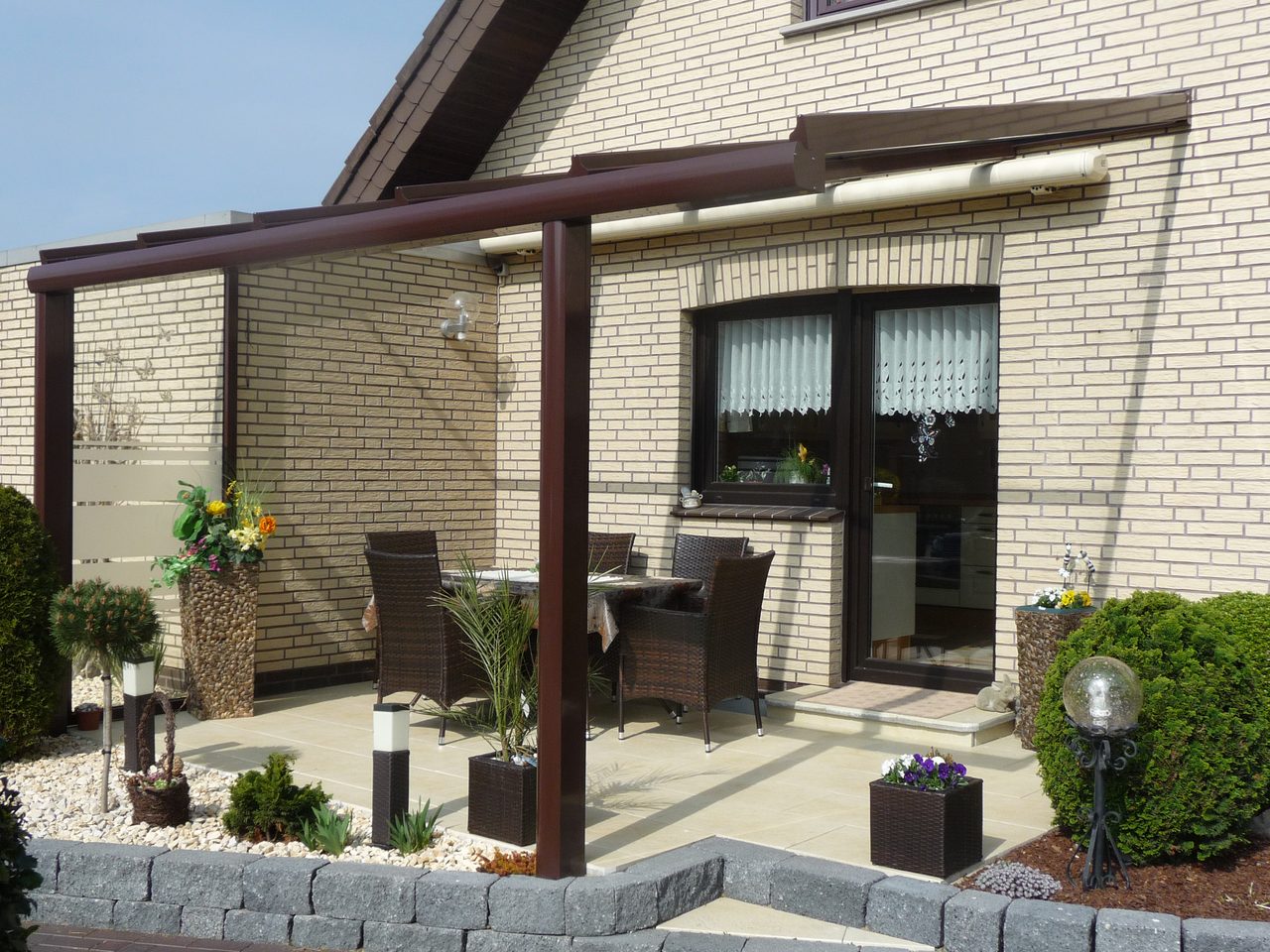 Dunkelbraune Terrassenüberdachung mit seitlichem Windfänger aus Glas an einem Klinkerhaus