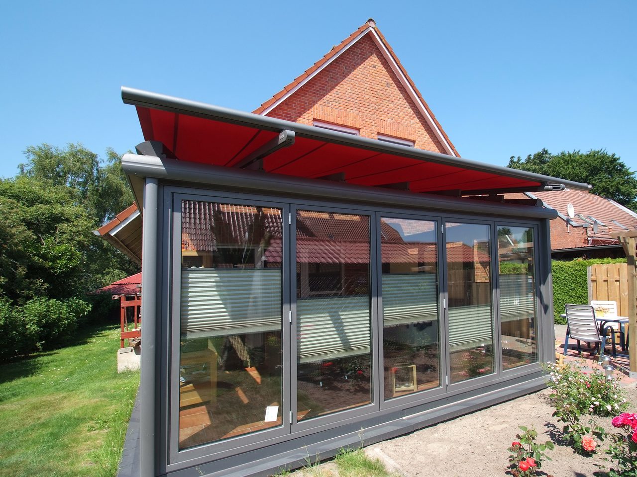 Grauer Wintergarten mit ausgefahrener roter Markise an einem Klinkerhaus