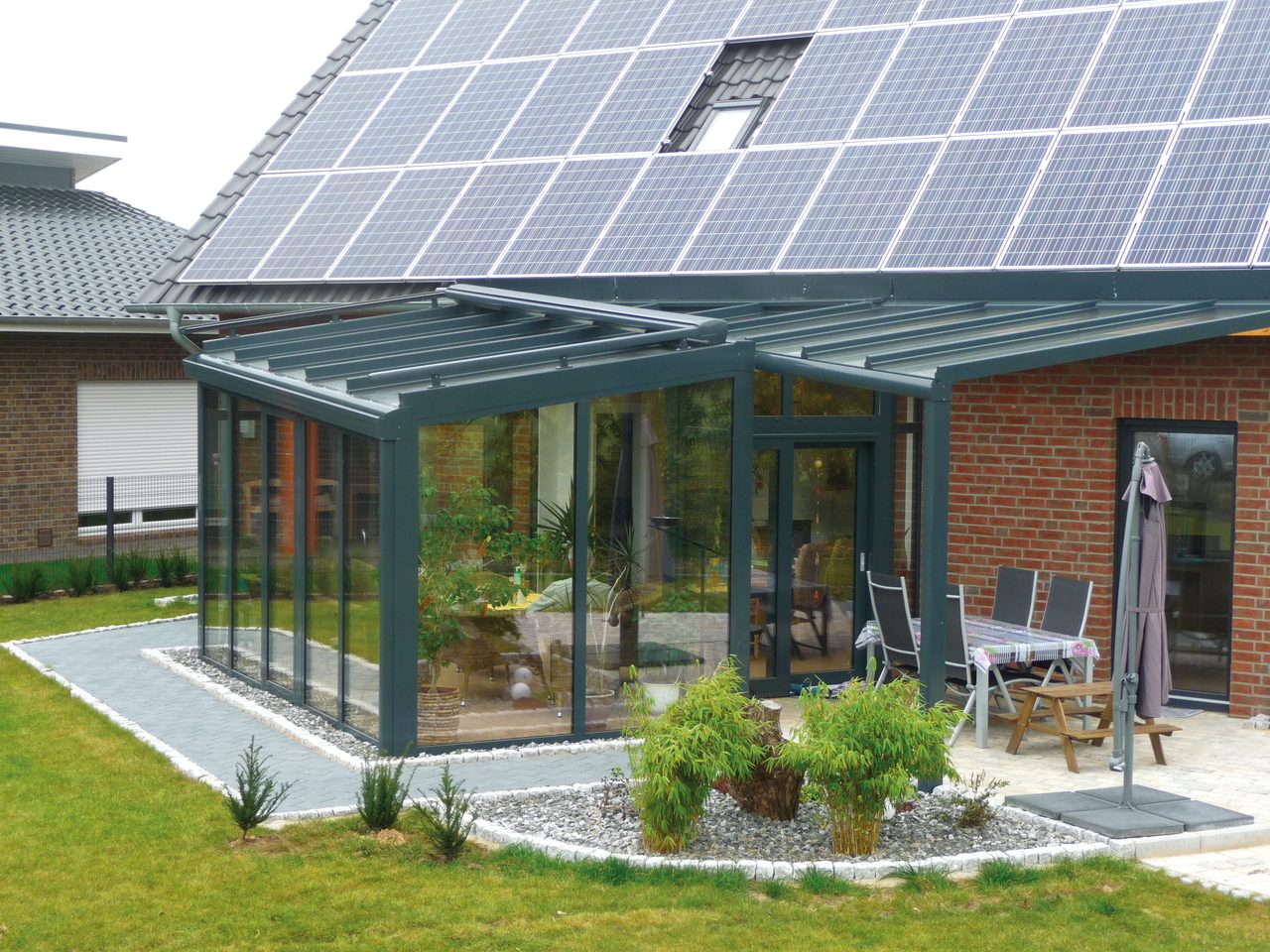 Anthrazitfarbener Wintergarten mit angrenzender Terrassenüberdachung an einem Klinkerhaus mit Solardach